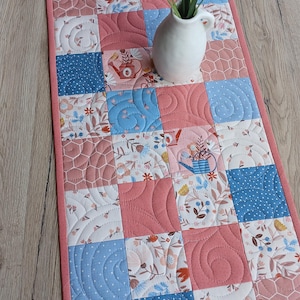 Può includere: Runner da tavolo trapuntato con un design patchwork. Il runner presenta quadrati nei toni del rosa, blu e bianco, con motivi floreali e geometrici. Un piccolo vaso bianco con fiori è posto sopra. Il runner è largo circa 30 cm.