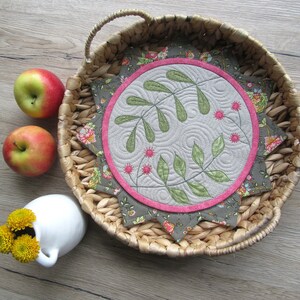 K&ouml;nnte beinhalten: Ein runder, gesteppter Untersetzer mit einem floralen Design in Gr&uuml;n-, Rosa- und Graut&ouml;nen. Der Untersetzer liegt auf einem geflochtenen Weidentablett.