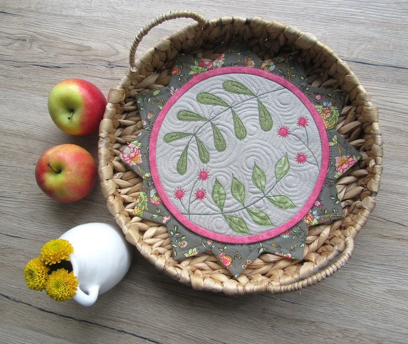 K&ouml;nnte beinhalten: Ein runder, gesteppter Untersetzer mit einem floralen Design in Gr&uuml;n-, Rosa- und Graut&ouml;nen. Der Untersetzer liegt auf einem geflochtenen Weidentablett.