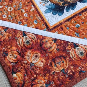 May include: A quilted fabric runner featuring a patchwork design with orange, brown, and white tones. The fabric displays repeating patterns of pumpkins, autumn leaves, and floral motifs, accented by intricate machine-stitched quilting lines.