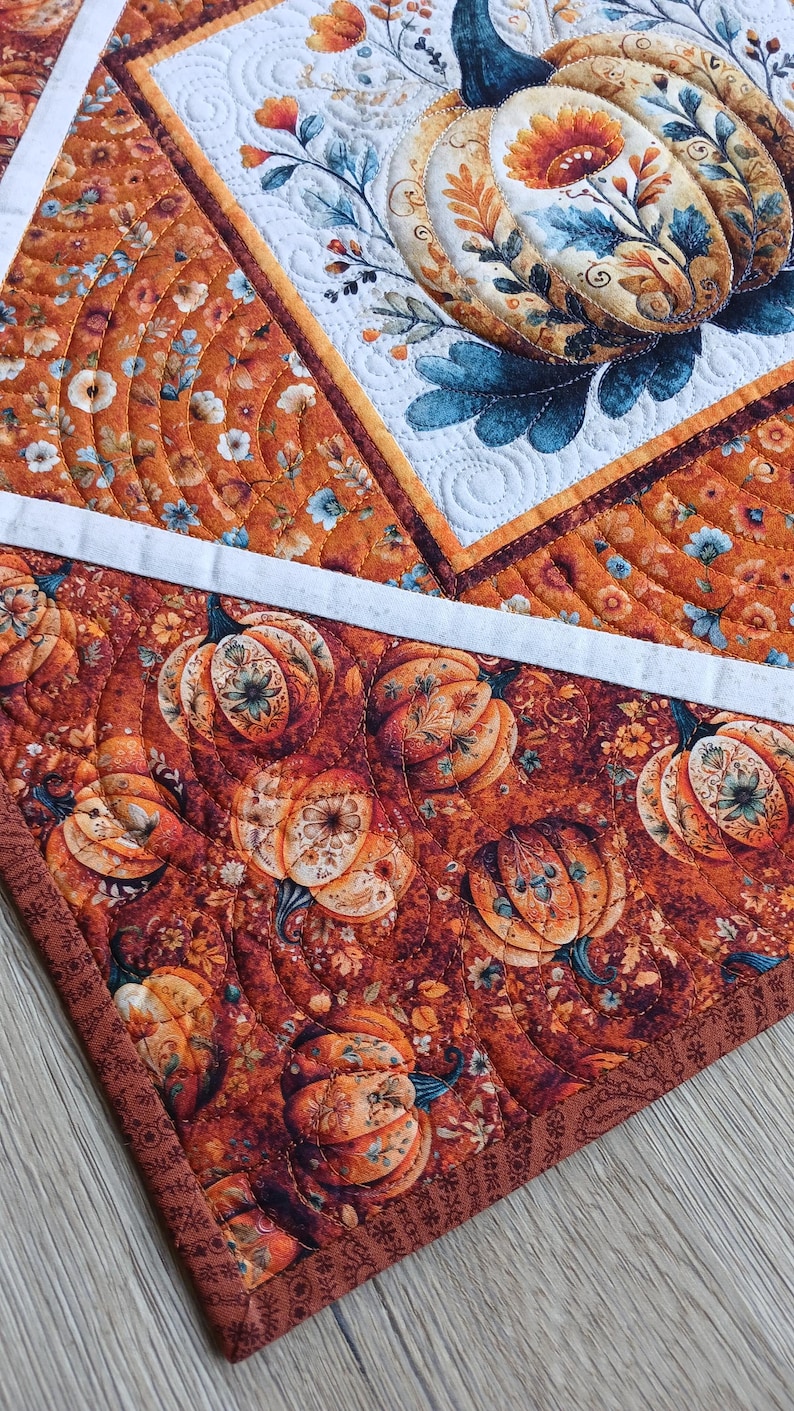 May include: A quilted fabric runner featuring a patchwork design with orange, brown, and white tones. The fabric displays repeating patterns of pumpkins, autumn leaves, and floral motifs, accented by intricate machine-stitched quilting lines.