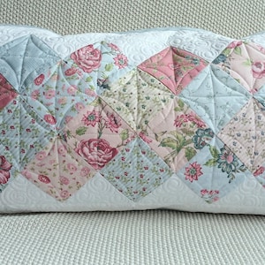Könnte beinhalten: Ein rechteckiges Zierkissen mit einem gesteppten Blumenmuster. Das Kissen hat ein Patchwork-Design mit rosa, blauen und weißen Blumenstoffquadraten. Die Kanten sind weiß mit einem gesteppten Muster.