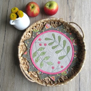 K&ouml;nnte beinhalten: Eine runde, gesteppte Tischunterlage mit einem floralen Design in den Farben Rosa, Gr&uuml;n und Braun. Die Unterlage ist von einem geflochtenen Weidenkorb umgeben.