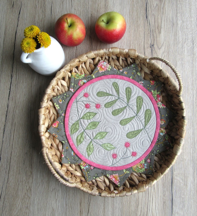 K&ouml;nnte beinhalten: Eine runde, gesteppte Tischunterlage mit einem floralen Design in den Farben Rosa, Gr&uuml;n und Braun. Die Unterlage ist von einem geflochtenen Weidenkorb umgeben.