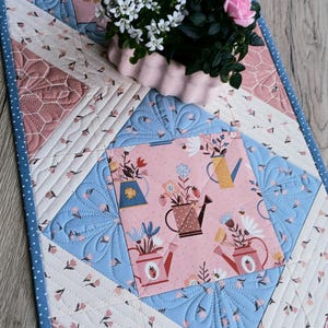 Könnte beinhalten: Gesteppter Tischläufer mit Blumen- und Gießkannenmuster in Rosa, Blau und Weiß. Der Läufer hat einen blauen Tupfenrand und einen rosa Blumentopf mit weißen und rosa Blumen.
