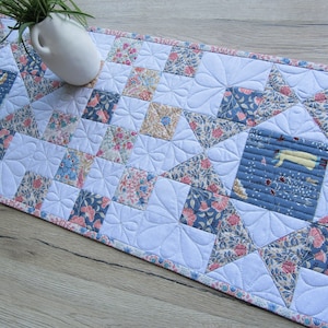 Könnte beinhalten: Ein weißer gesteppter Tischläufer mit Blumenmuster. Der Läufer besteht aus Stoffquadraten, die in einem Patchwork-Design zusammengenäht sind. Der Läufer hat einen gewellten Rand und ist mit einem zarten Blumenmuster verziert.