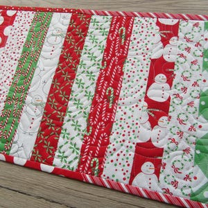 May include: A festive Christmas table runner with a patchwork design. It features alternating vertical stripes of red, white, and green fabrics. Patterns include snowmen, candy canes, polka dots, and snowflakes. The edges are trimmed with a red and white striped border.