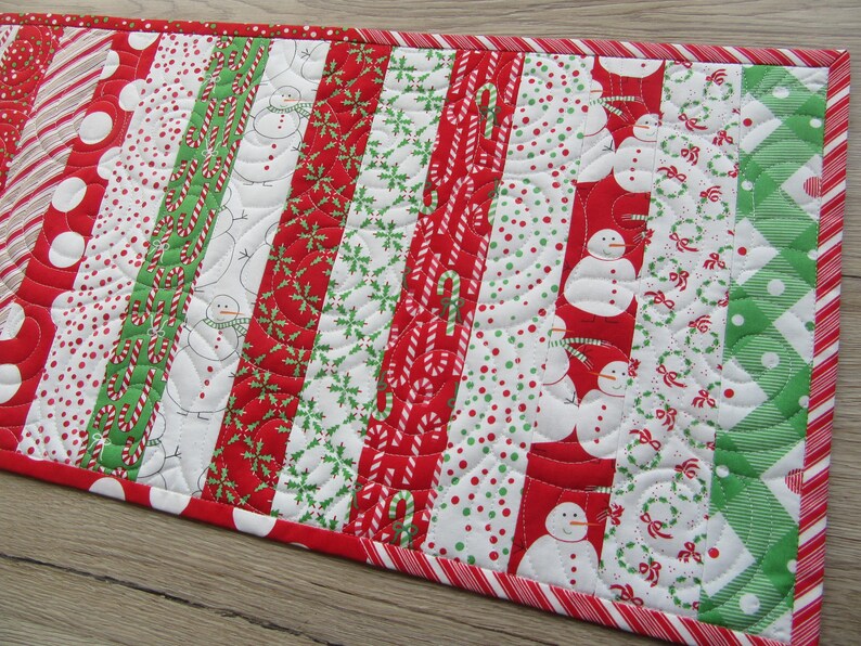 May include: A festive Christmas table runner with a patchwork design. It features alternating vertical stripes of red, white, and green fabrics. Patterns include snowmen, candy canes, polka dots, and snowflakes. The edges are trimmed with a red and white striped border.