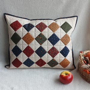 May include: A rectangular quilted pillow featuring a cream-colored base with a grid of diamond-shaped fabric patches in various colors including red, navy blue, olive green, and mustard yellow, all finished with a dark navy blue border.