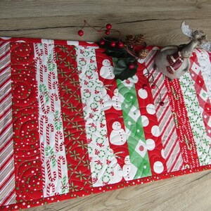 May include: A festive Christmas table runner with a patchwork design. The runner features alternating stripes of red, white, and green patterns, including candy canes, snowmen, and polka dots. A decorative reindeer figurine and berries are on top.