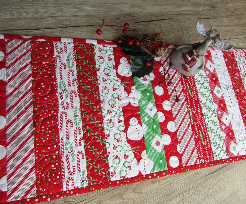 May include: A festive Christmas table runner with a patchwork design. The runner features alternating stripes of red, white, and green patterns, including candy canes, snowmen, and polka dots. A decorative reindeer figurine and berries are on top.