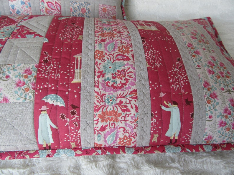May include: A red and white patterned pillow with a gray linen border. The pillow is quilted with a floral design and features a small figure holding an umbrella.