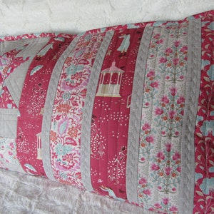 May include: A long, rectangular pillow with a patchwork design. The pillow is made of various fabrics, including a red floral print, a gray linen, and a white floral print. The pillow is quilted with a simple stitch pattern.