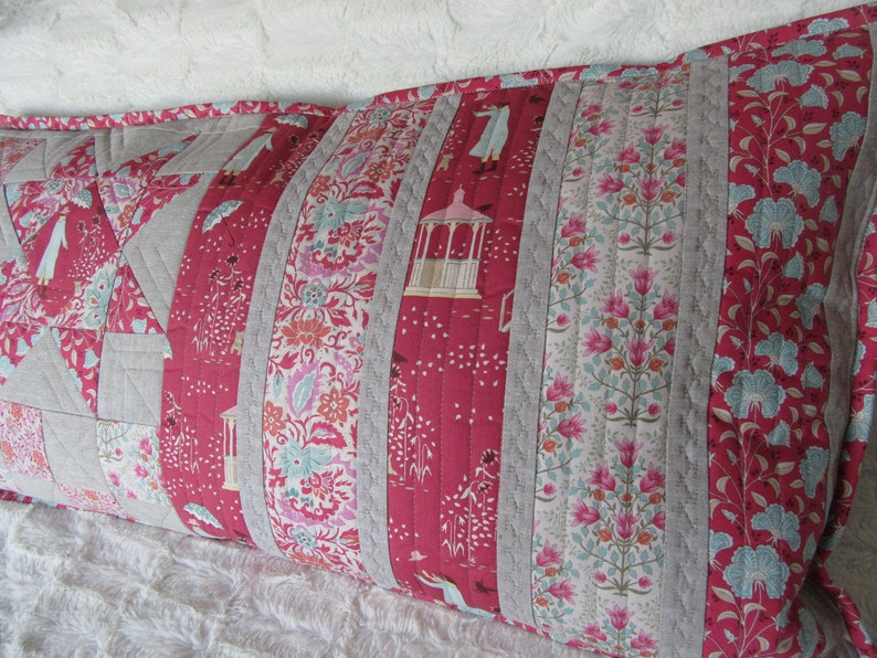May include: A long, rectangular pillow with a patchwork design. The pillow is made of various fabrics, including a red floral print, a gray linen, and a white floral print. The pillow is quilted with a simple stitch pattern.