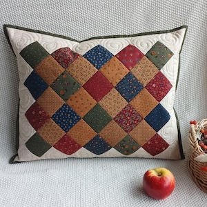 May include: A square throw pillow featuring a patchwork design of diamond-shaped fabric pieces in various colors including red, blue, green, and gold. The pillow has a cream-colored background with quilted swirl patterns and a dark green border.