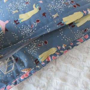 May include: A blue pillow with a zipper opening. The pillow fabric has a repeating pattern of people walking in a park with trees and flowers. The pillow is lying on a white fluffy surface.