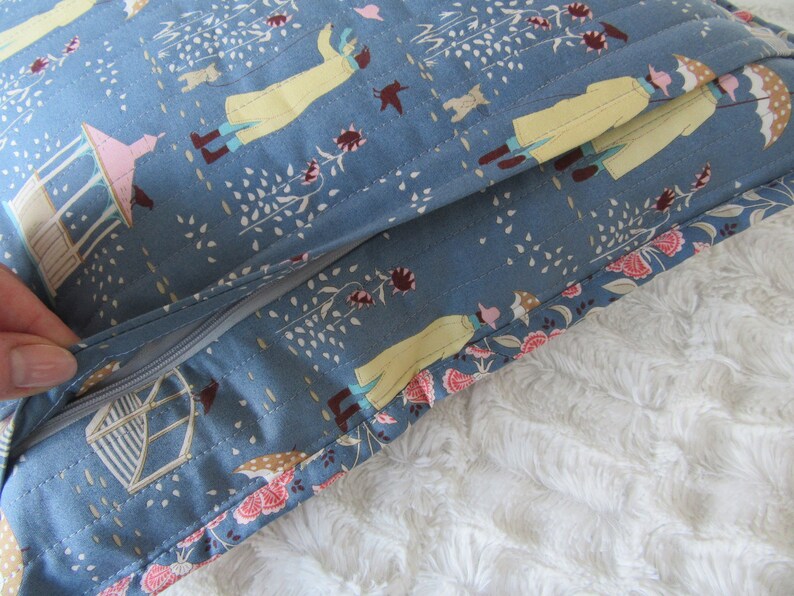 May include: A blue pillow with a zipper opening. The pillow fabric has a repeating pattern of people walking in a park with trees and flowers. The pillow is lying on a white fluffy surface.
