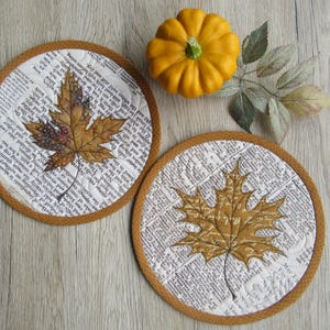 Könnte beinhalten: Zwei runde, gesteppte Untersetzer mit Herbstthema. Jeder Untersetzer zeigt ein gedrucktes Ahornblatt-Design in Braun- und Goldtönen, vor einem Hintergrund aus gedrucktem Text. Ein kleiner oranger Kürbis und Herbstblätter vervollständigen die herbstliche Anordnung.