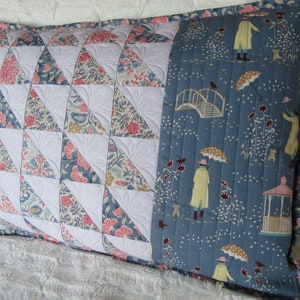 May include: A blue and white patterned pillow with a quilted design. The pillow has a blue fabric panel with a white bridge, umbrellas, and people walking in the rain.