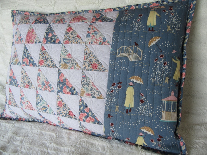 May include: A blue and white patterned pillow with a quilted design. The pillow has a blue fabric panel with a white bridge, umbrellas, and people walking in the rain.