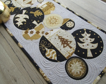 May include: A quilted Christmas table runner featuring large, circular ornaments in black, gold, and cream. The ornaments are decorated with stars, trees, and other festive designs. The runner is approximately 12 inches wide and has a gray background with a swirling pattern.