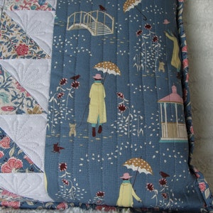 May include: A blue quilted pillow with a white and floral patterned fabric on the back. The front of the pillow features a repeating pattern of umbrellas, birds, and a gazebo.