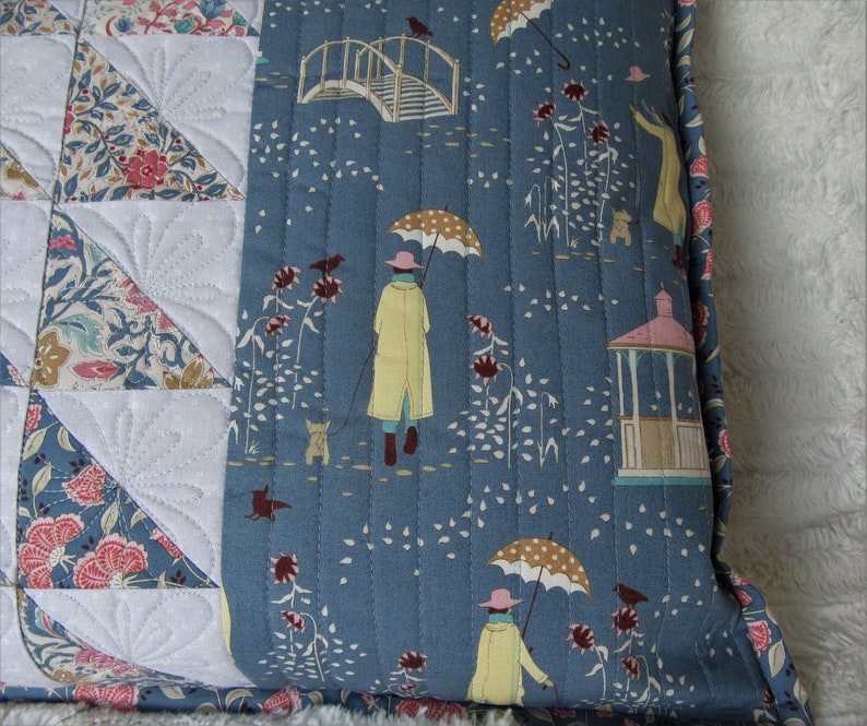 May include: A blue quilted pillow with a white and floral patterned fabric on the back. The front of the pillow features a repeating pattern of umbrellas, birds, and a gazebo.