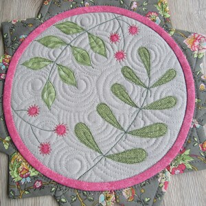 K&ouml;nnte beinhalten: Ein runder, grauer Stoffuntersetzer mit rosa Rand und floralem Muster. Die Mitte des Untersetzers zeigt ein gr&uuml;nes gesticktes Design aus Bl&auml;ttern und Beeren.