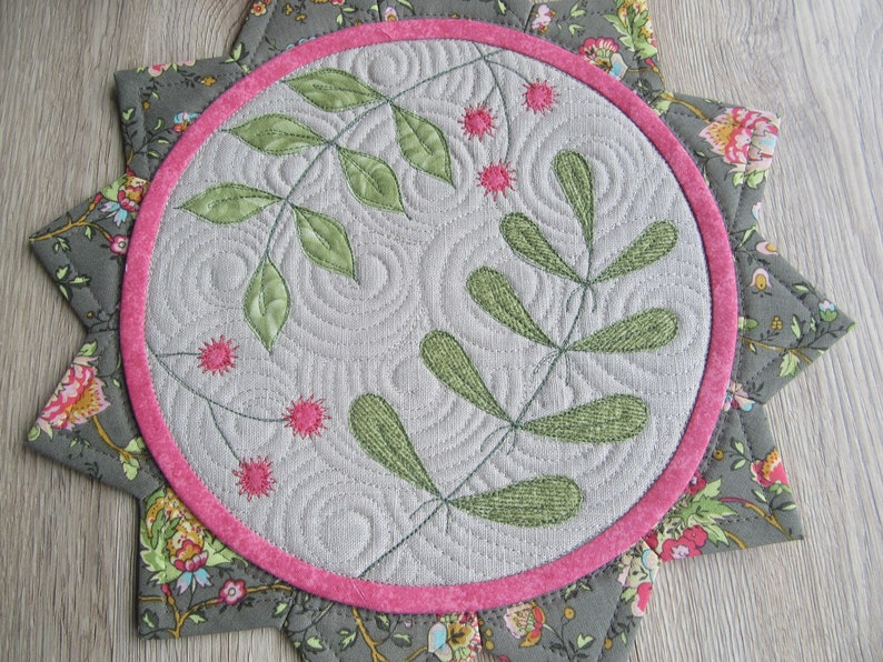 K&ouml;nnte beinhalten: Ein runder, grauer Stoffuntersetzer mit rosa Rand und floralem Muster. Die Mitte des Untersetzers zeigt ein gr&uuml;nes gesticktes Design aus Bl&auml;ttern und Beeren.