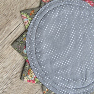 K&ouml;nnte beinhalten: Ein runder Platzteller in Grau mit wei&szlig;en Punkten und einem floralen Muster am Rand. Der Platzteller ist aus Stoff und hat eine sternf&ouml;rmige Form.