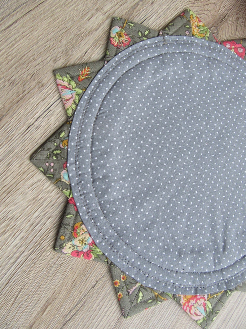 K&ouml;nnte beinhalten: Ein runder Platzteller in Grau mit wei&szlig;en Punkten und einem floralen Muster am Rand. Der Platzteller ist aus Stoff und hat eine sternf&ouml;rmige Form.