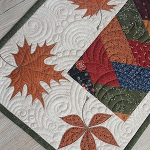 May include: A rectangular fabric table runner featuring a cream-colored background with quilted swirl patterns and three appliqued orange maple leaves. One side displays a patchwork section with floral and geometric prints in red, green, and blue.
