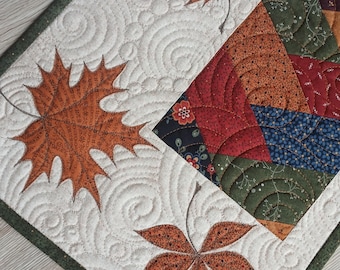 May include: A rectangular fabric table runner featuring a cream-colored background with quilted swirl patterns and three appliqued orange maple leaves. One side displays a patchwork section with floral and geometric prints in red, green, and blue.