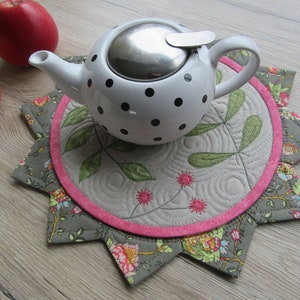 K&ouml;nnte beinhalten: Eine wei&szlig;e Teekanne mit schwarzen Punkten und einem silbernen Deckel steht auf einem grauen und rosafarbenen gesteppten Untersetzer mit Blumenmuster.