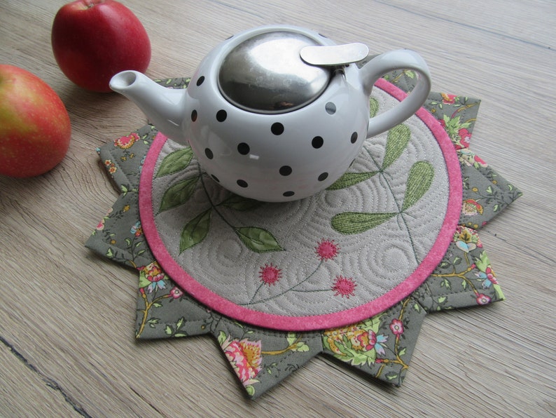 K&ouml;nnte beinhalten: Eine wei&szlig;e Teekanne mit schwarzen Punkten und einem silbernen Deckel steht auf einem grauen und rosafarbenen gesteppten Untersetzer mit Blumenmuster.