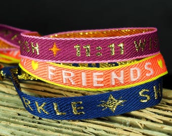 Op de afbeelding: Stapel van drie geweven vriendschapsarmbanden in blauwe, oranje en paarse tinten. De armbanden hebben gouden tekst met de tekst "SPARKLE", "FRIENDS" en "11:11 WISH". Elke armband heeft een gouden ster of gouden accenten.