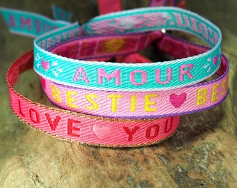 Op de afbeelding: Drie kleurrijke vriendschapsarmbanden met de woorden "Amour", "Bestie" en "Love You" geweven in de stof.