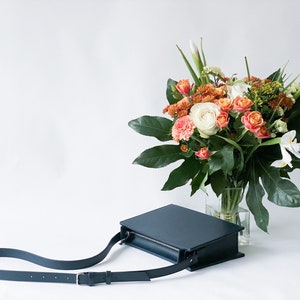 May include: A dark blue leather crossbody bag with a long strap. The bag is sitting on a white surface next to a bouquet of flowers in a clear glass vase.