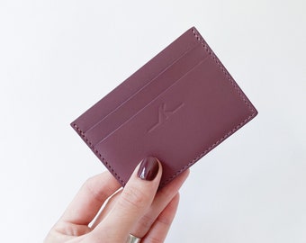 May include: A burgundy leather card holder with three card slots and a debossed logo. The rectangular wallet is held against a white background. The card holder is a deep reddish-purple color.