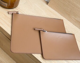 May include: Two tan leather pouches of different sizes are displayed. The larger pouch is rectangular, with a zipper closure and a small logo in the upper right corner. The smaller pouch is placed in front of the larger one. Both pouches have a smooth, clean design.