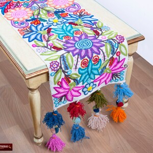 May include: White table runner with colorful embroidered floral design and tassels. The runner features a variety of flowers in shades of pink, purple, blue, green, and red.