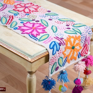 May include: A white table runner with colorful embroidered flowers and green leaves. The runner has a fringe with colorful pom-poms.