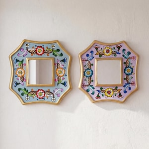 May include: Two decorative wall mirrors with floral designs. One mirror has a light blue background, the other is lavender. Both have gold trim and square mirrors. The floral patterns include red, orange, yellow, and blue flowers.