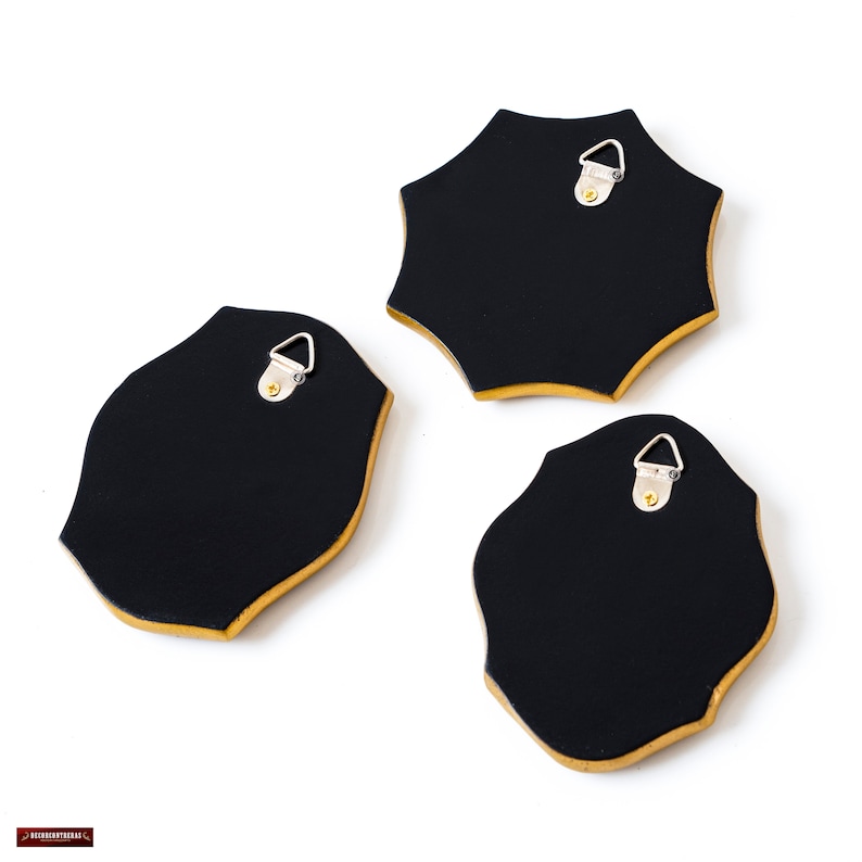 May include: Three black wall decor pieces with gold trim and a unique shape. Each piece has a metal hanger on the back for easy installation. The pieces are set against a white background.