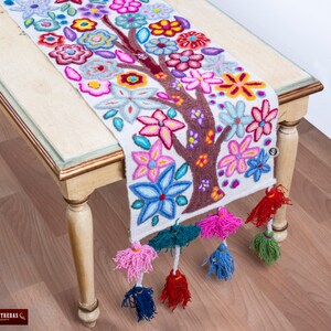 May include: A white table runner with a colorful embroidered tree design. The tree is surrounded by flowers in shades of pink, blue, yellow, and red. The runner has tassels on the ends.