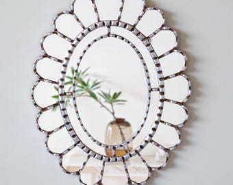 Op de afbeelding: Ovale spiegel met een geschulpte rand. De spiegel heeft een zilveren frame met een decoratief patroon en een centrale ovale spiegel. De spiegel reflecteert een vaas met groen.