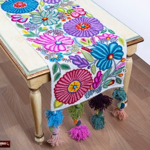 May include: A white table runner with colourful embroidered flowers. The runner has tassels on the ends and is decorated with pink, purple, blue, green, and yellow flowers.