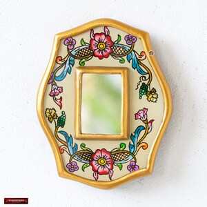 May include: Decorative wall mirror with a gold-painted frame and a central rectangular mirror. The frame features a cream-colored background with a floral design in red, pink, blue, and green. The mirror is an oval shape.