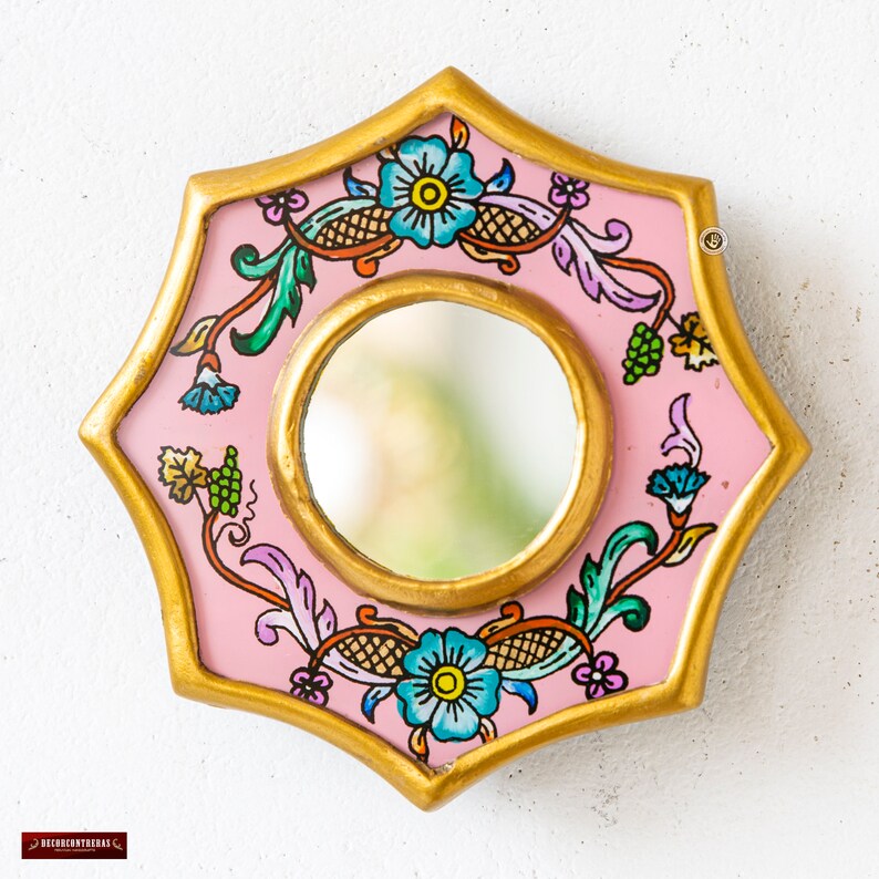 May include: A decorative, star-shaped mirror with a pink frame and a gold border. The mirror features hand-painted floral and vine designs in blue, green, and purple. The central mirror is round and has a gold frame.