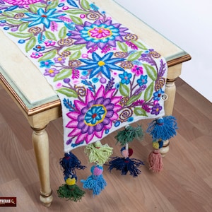 May include: White table runner with colorful embroidered flowers and tassels. The runner features a floral design with pink, purple, blue, and green flowers. The tassels are blue, green, and pink.